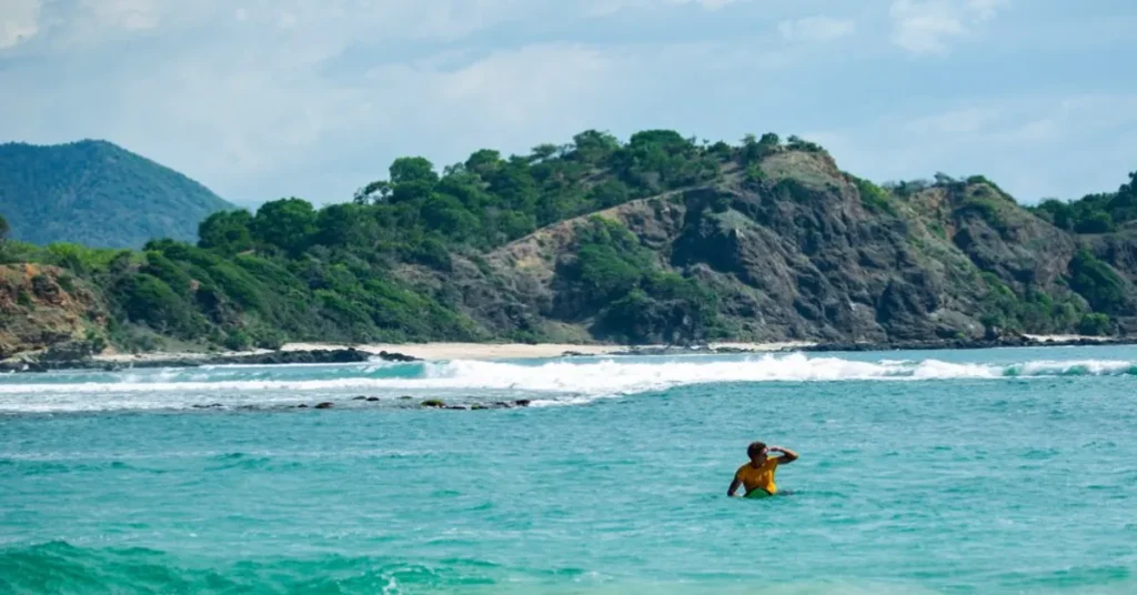 A person standing in clear turquoise water near a lush green island with rocky cliffs in Indonesia, representing a serene beach getaway.